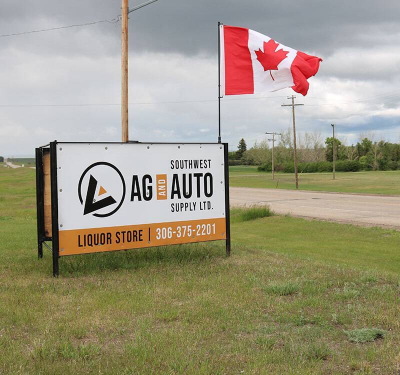 Southwest Ag & Auto store sign outside in green grass with paved road and Canadian flag hanging behind it
